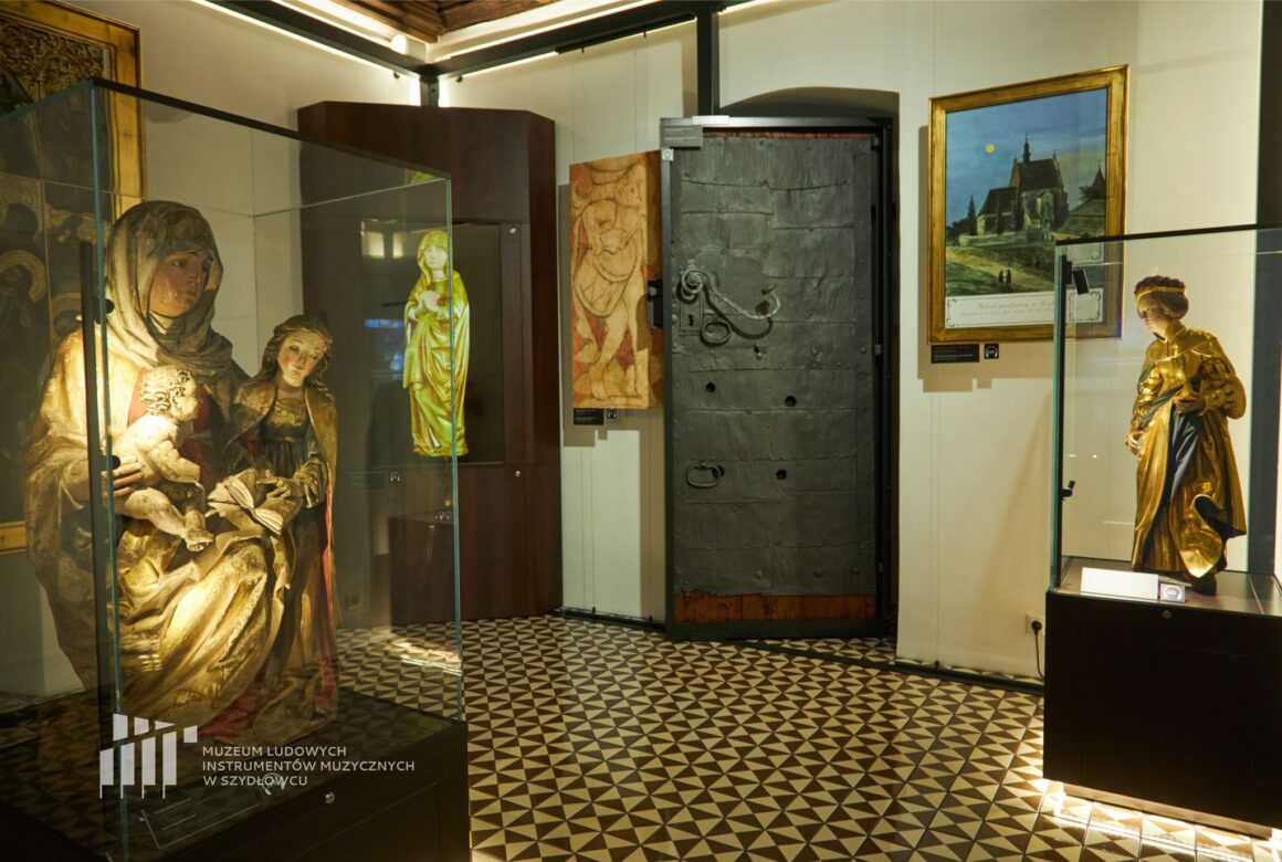 Fragment wystawy Zamek w Szydłowcu. Po lewej stronie rzeźba św. Anny. Po prawej złota rzeźba św. Barbary. W tle metalowe drzwi i hologram.