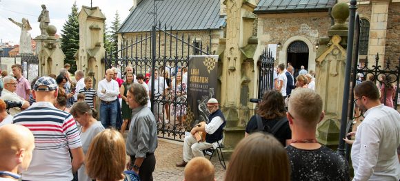 Na zdjęciu ludzie wychodzący z kościoła. Pomiędzy nimi siedzi mężczyzna i gra na lirze.