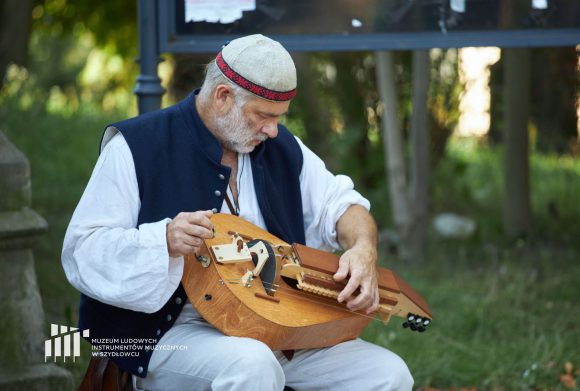 Mężczyzna w białej koszuli i granatowej kamizelce siedzi. Na kolanach trzyma instrument muzyczny. Gra na nim.