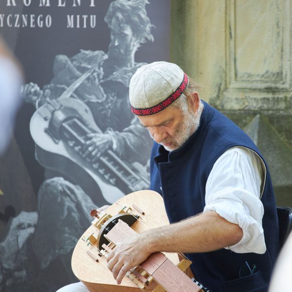 Na tle kamiennych murów siedzi mężczyzna i gra na lirze korbowej.