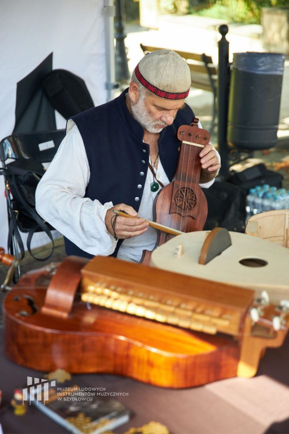 Na środku zdjęcia siedzi mężczyzna i gra na instrumencie smyczkowym. W dolnej części zdjęcia leżą liry korbowe.