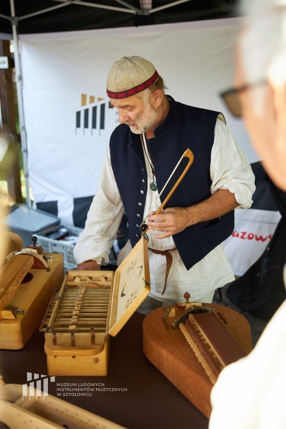 na środku zdjęcia mężczyzna z brodą. Przed nim leżą drewniane instrumenty muzyczne.