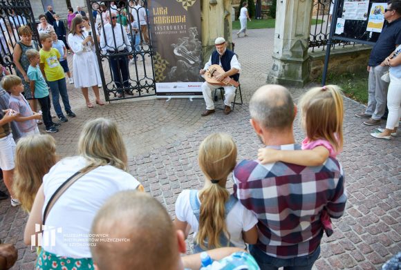 Grupa ludzi stojąca w kole słucha muzyki grajka.