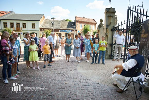 Duża grupa ludzi stoi i słucha mężczyzny grającego na lirze.