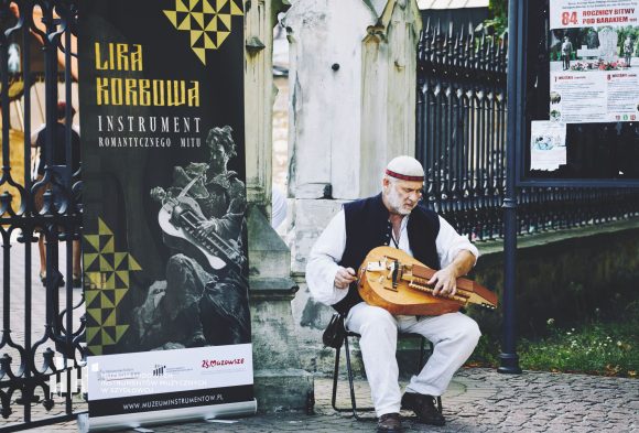 Mężczyzna z brodą siedzi przed wysokim ogrodzeniem i gra na lirze.