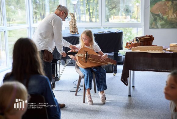 Dziewczynka z długimi włosami siedzi na krześle i trzyma duży instrument muzyczny. Mężczyzna w białej koszuli coś jej pokazuje.