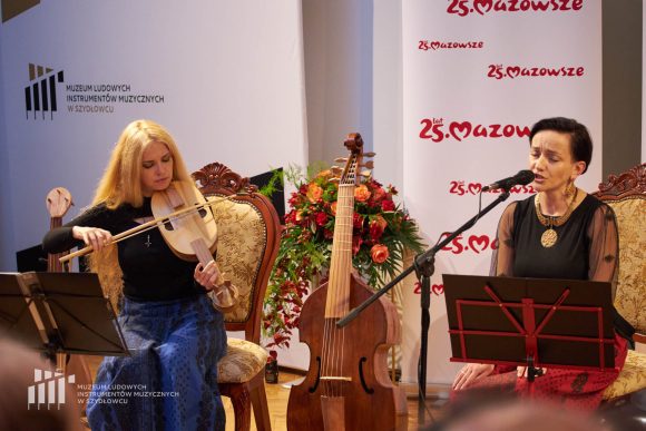 Zdjęcie. Dwie kobiety siedzą na krzesłach na scenie. Kobieta po prawej stronie śpiewa do mikrofonu. Kobieta po lewej gra na instrumencie.