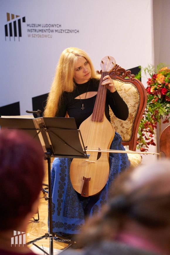 Zdjęcie. Kobieta z długimi blond włosami siedzi na krześle. Trzyma instrument przypominający basy. Zespół Wernyhora.