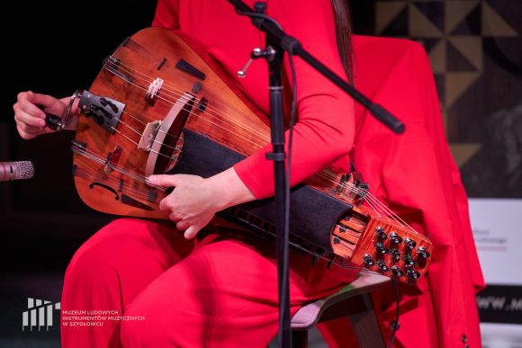 W centralnej części zdjęcia znajduje się duży drewniany instrument muzyczny - lira korbowa. trzyma go osoba ubrana na czerwono.