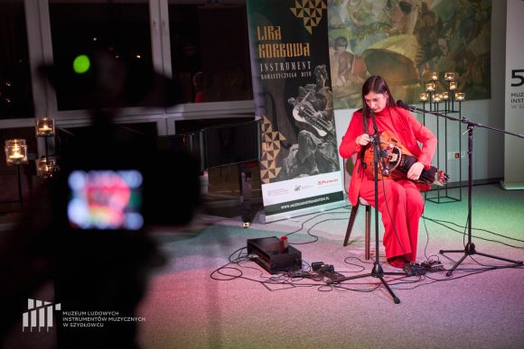 Z lewej strony zdjęcia cień kamery. Na środku kobieta grająca lirze. Na podłodze kable i mikrofony. Z tyłu świecznik.