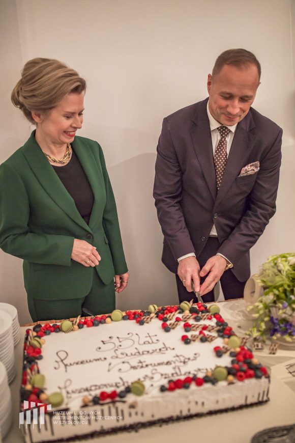 Kobieta w zielonym garniturze i mężczyzna w szarej marynarce kroją tort.