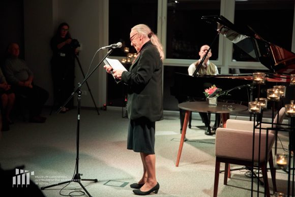 W centralnej części stoi kobieta, która czyta do mikrofonu. W tle po prawej stronie mężczyzna siedzi za czarnym fortepianem.