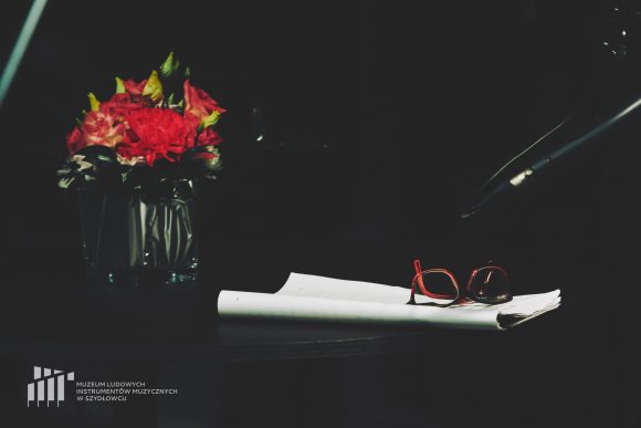 Po lewej stronie zdjęcia na czarnym tle niski wazon z różowymi kwiatami. Po prawej stronie na białych kartkach leżą okulary.