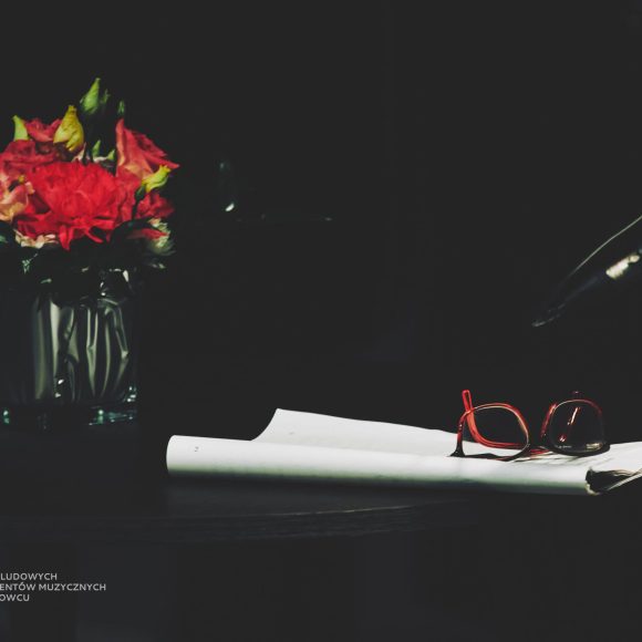 Po lewej stronie zdjęcia na czarnym tle niski wazon z różowymi kwiatami. Po prawej stronie na białych kartkach leżą okulary.