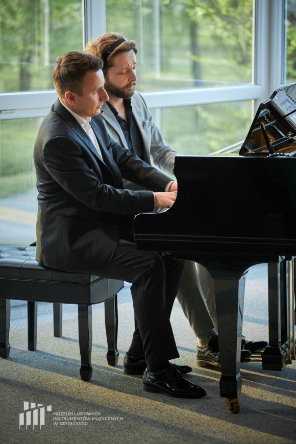 Dwóch mężczyzn siedzących i grających na fortepianie na tle dużych okien.