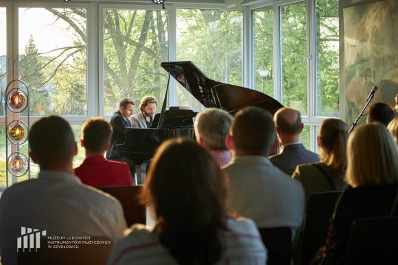 Dwóch mężczyzn siedzących i grających na fortepianie na tle dużych okien przed publicznością siedzącą na krzesłach.