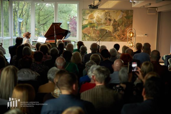 Publiczność w sali z oknami, przed którą siedzą i grają dwaj mężczyźni na czarnym fortepianie.