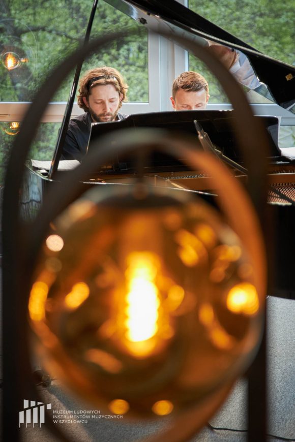 Dwaj mężczyźni za rozmytym zbliżeniem okrągłej lampy siedzą za czarnym fortepianem.