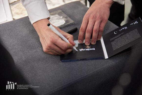 Dłonie podpisujące czarną okładkę płyty białym markerem na stoliku z ciemnym obrusem.