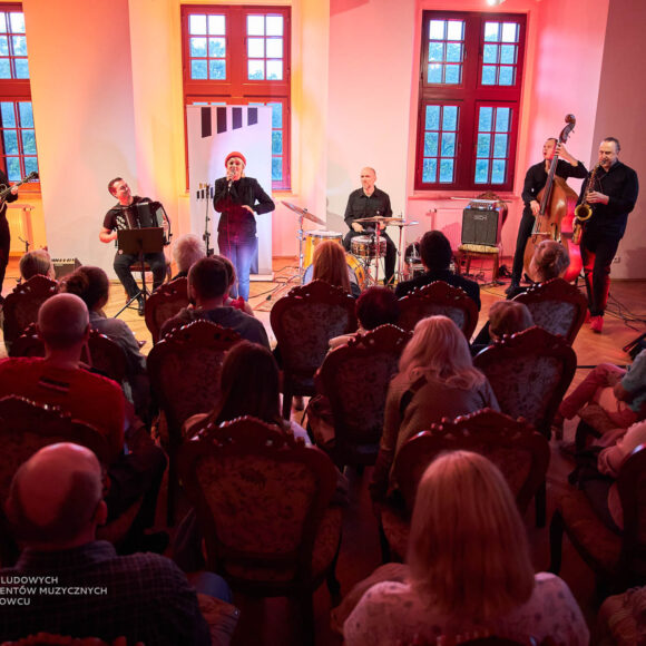 Publiczność siedząca na krzesłach tyłem przed zespołem pięciu mężczyzn, którzy trzymają instrumenty muzyczne i jednej kobiety która trzyma mikrofon.