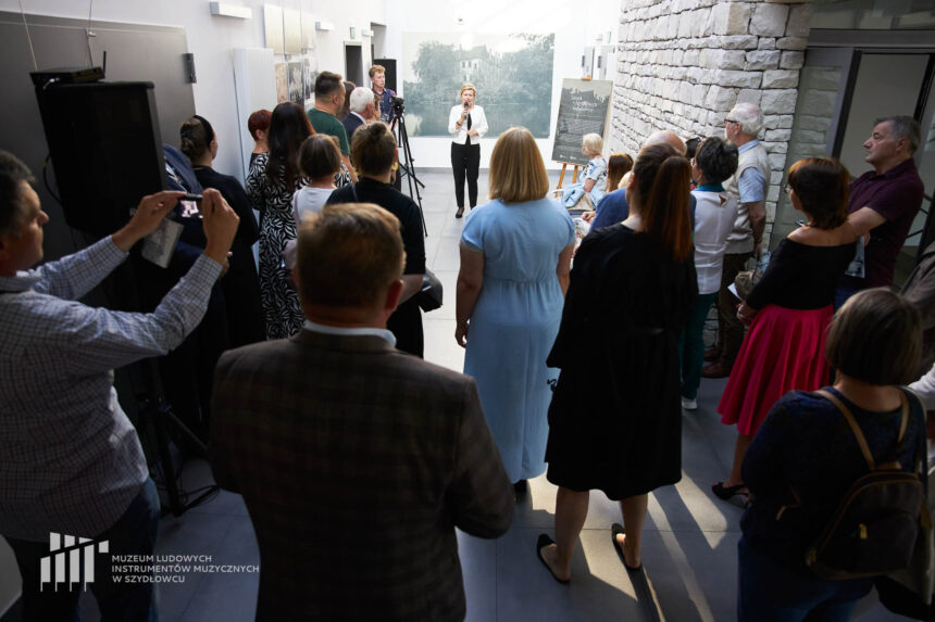 Grupa osób stojących tyłem patrzących na kobietę trzymającą mikrofona na tle ściany z dużym zdjęciem zniszczonego zamku.