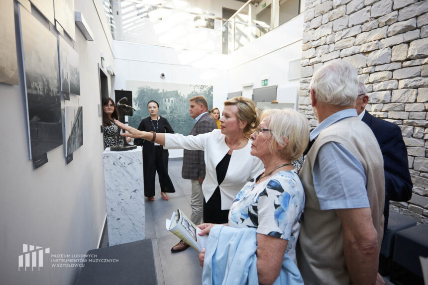 Kobieta pokazująca zdjęcia zawieszone na ścianie grupie osób oglądających wystawę fotografii przedstawiających zamek.