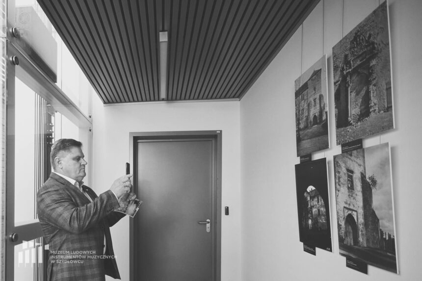 Czarno-białe zdjęcie przedstawiające mężczyznę stojącego na korytarzu i robiącego zdjęcie przy pomocy telefony komórkowego wiszącym na ścianie oprawionym w szklaną antyramę fotografiom.