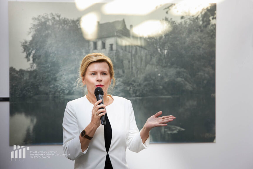 Kobieta mówiąca do mikrofonu z uniesiąną lewą otwartą ku górze dłonią na tle zdjęcia przedstawiającego zniszczony zamek wśród drzew.