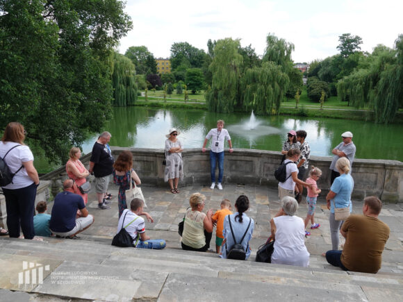 Grupa osób stojąca na schodach kamiennych z balustradą z widokiem na wodę w fosie i park z drzewami.