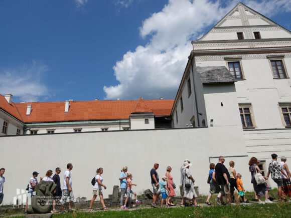 Grupa ludzi idąca po chodniku z kamienia przy murach i fragmencie zamku w Szydłowcu