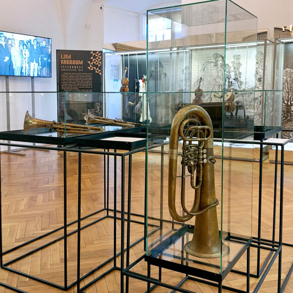 blaszane instrumenty muzyczne umieszczone w szklanych gablotach ustawionych na czarnych metalowych nogach, z prawej strony kartka z opisami umieszczona na stelażu zabudowy.