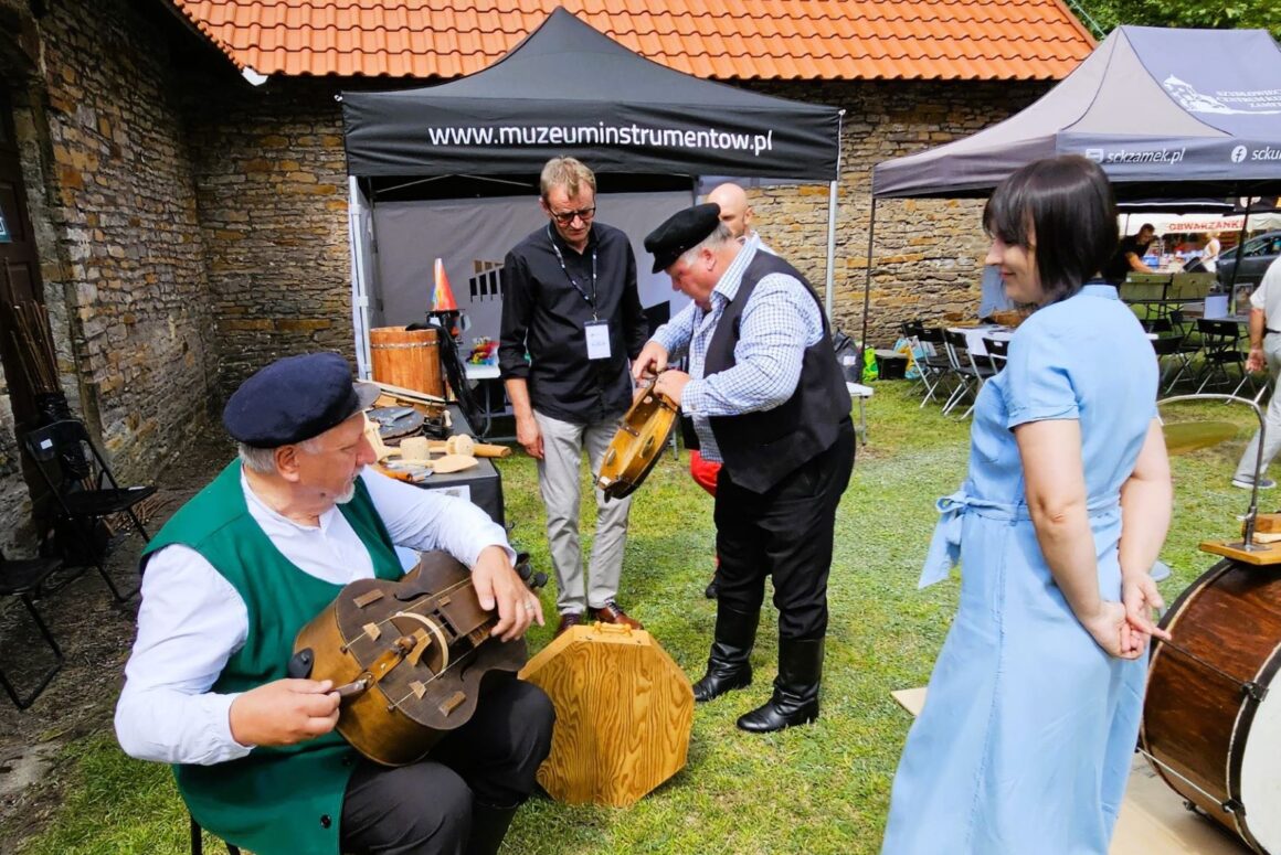 Siedzący mężczyzna z instrumentem na kolanach ubrany w strój ludowy w zieloną kamizelkę i czapkę, obopk niego stoi kobieta w długiej sukience a za nimi trzy osoby oglądają drewniane instrumenty muzyczne