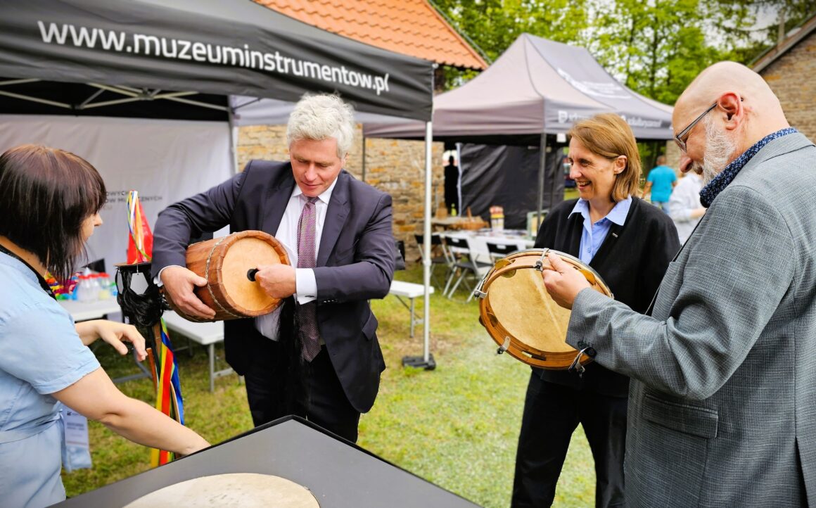 Cztery dorosłe stojące osoby na trawniku przed namiotami plenerowymi przy czarnym stoliku. Dwaj starsi mężczyźni trzymają okrągłe instrumenty muzyczne.