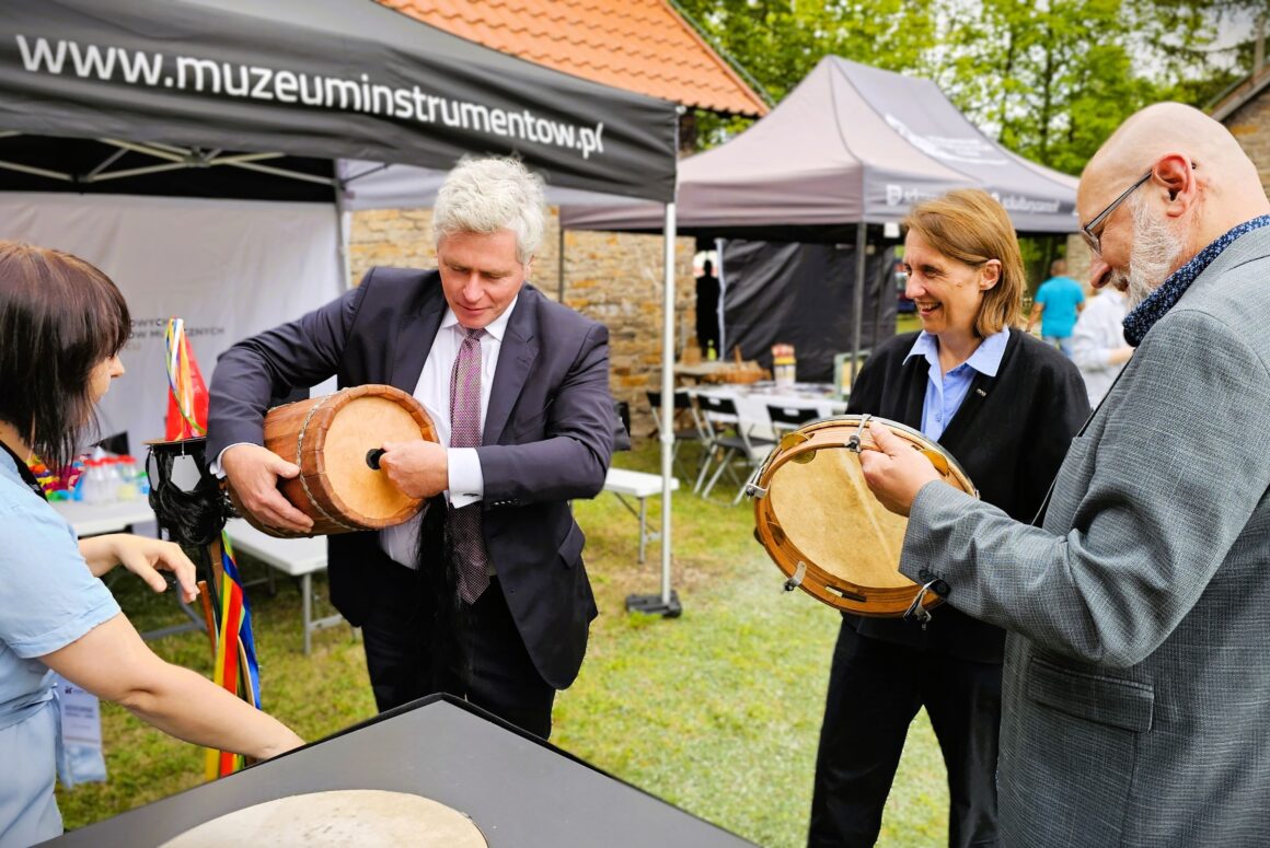 Cztery dorosłe stojące osoby na trawniku przed namiotami plenerowymi przy czarnym stoliku. Dwaj starsi mężczyźni trzymają okrągłe instrumenty muzyczne.