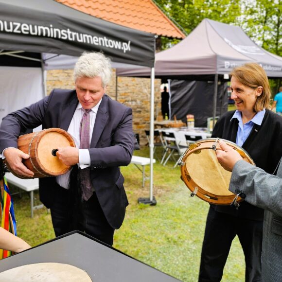 Cztery dorosłe stojące osoby na trawniku przed namiotami plenerowymi przy czarnym stoliku. Dwaj starsi mężczyźni trzymają okrągłe instrumenty muzyczne.