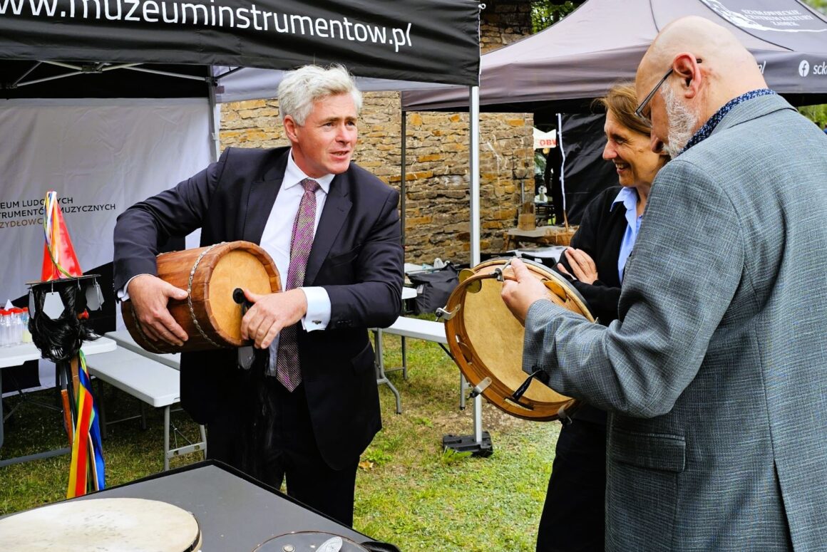 Trzy osoby stojące za czarnym stolikiem na którym poukładane są drewniane małe instrumenty muzyczne i ulotki. Mężczyźni trzymają instrumenty i rozmawiają. Za nimi czarny namiot z napisem www.muzeuminstrumentow.pl