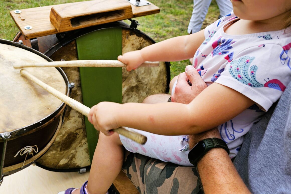 Dziecko widoczne od brody do dołu siedzące na kolanach dorosłej osoby. Dziecko trzyma pałeczki drtewniane w obu rękach i gra na ludowej perkusji.