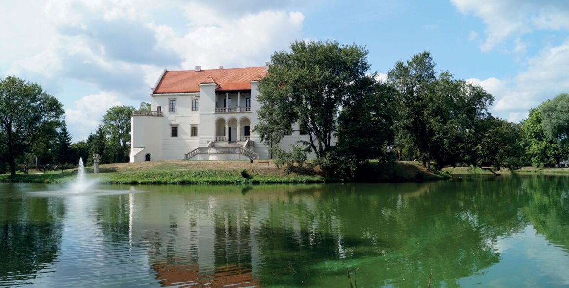 Biały zamek za fosą na której tryska fontanna. Zamek w otoczeniu wysokich drzew z czerwonym dachem.