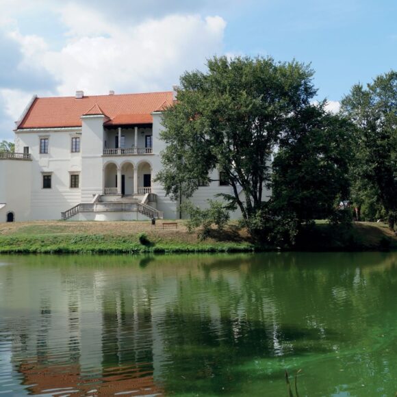 Biały zamek za fosą na której tryska fontanna. Zamek w otoczeniu wysokich drzew z czerwonym dachem.