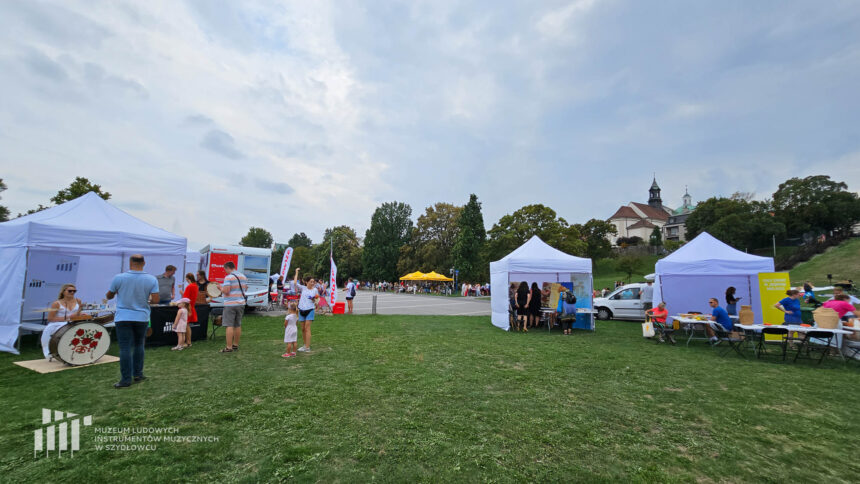 Przestrzeń z zielonym trawnikiem na którym ustawione są białe namioty firmowe ze stolikami przy których stoją ludzie. Za namiotami asfaltowe boisko a za drzewami u góry wieża kościoła.