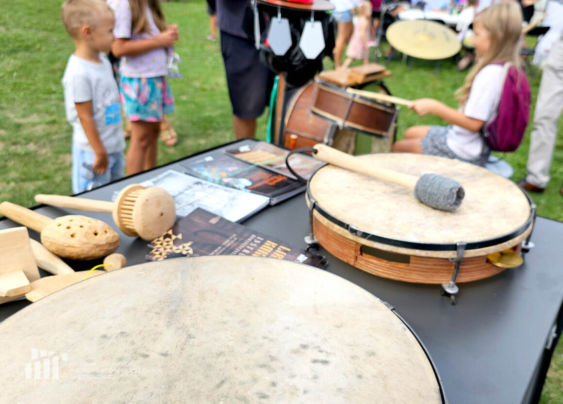 Drewniane instrumenty i narzeędzia muzyczne leżące na czarnym blacie stolika razem z ulotkami. Z tyłu w tle rozmyci ludzie grający na perkusji ludowej.