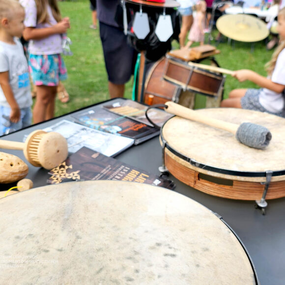 Drewniane instrumenty i narzeędzia muzyczne leżące na czarnym blacie stolika razem z ulotkami. Z tyłu w tle rozmyci ludzie grający na perkusji ludowej.