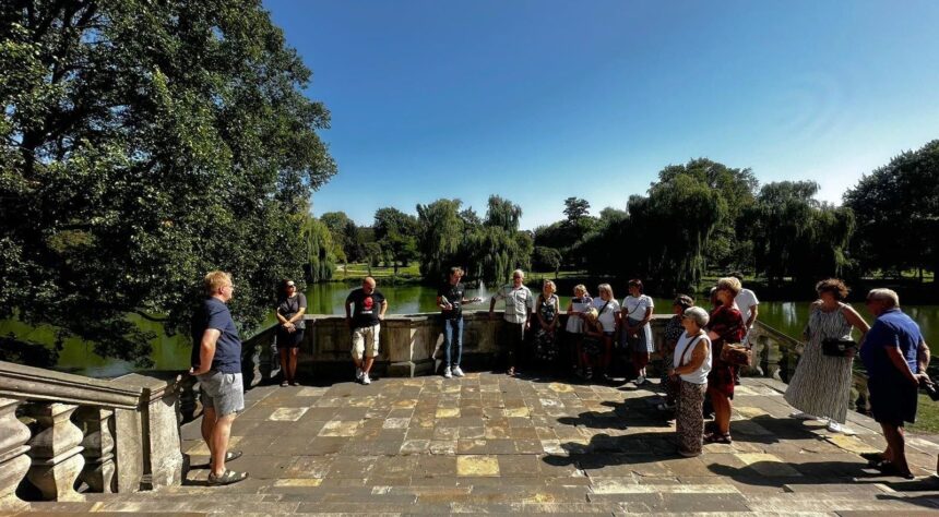 Grupa ludzi stojących na kamiennym balkonie z balustradą i widokiem na park i fosę z fontanną.