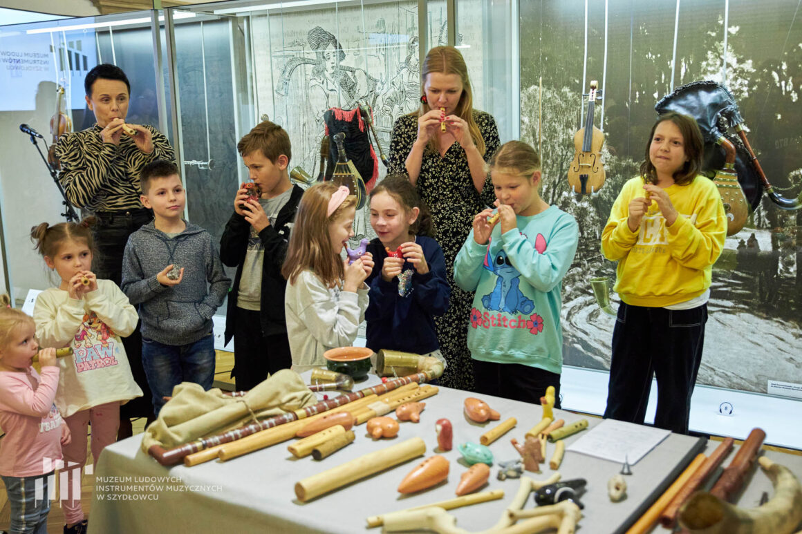 Dwie dorosłe kobiety i ośmioro dzieci trzymają w dłoniach i dmuchają w drewniane rurki i ceramiczne zwierzątka. Przed nimi stoi stolik z instrumentami muzycznymi. W tle szklana gablota z instrumentami muzycznymi.