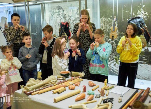 Dwie dorosłe kobiety i ośmioro dzieci trzymają w dłoniach i dmuchają w drewniane rurki i ceramiczne zwierzątka. Przed nimi stoi stolik z instrumentami muzycznymi. W tle szklana gablota z instrumentami muzycznymi.