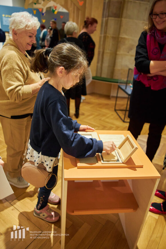 Dziewczynka w granatowej bluzce z torebeczką na ramieniu stoi przy stoliku i odbija pieczątkę. Za nią stoją dorosłe kobiety i patrzą na nią, w tle sala muzealna z ludźmi.