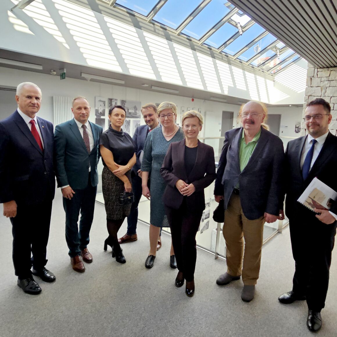 Osiem osób ubranych w garnitury i eleganckie stroje stojących w lobby z szarymi ścianami ze zdjęciami na ścianach i przeszklonym dachem.