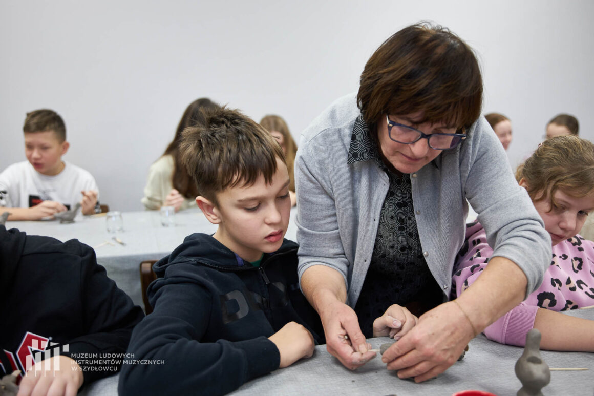 Kobieta z ciemnymi krótkimi włosami w okularach pokazuje chłopcu ubranemu w ciemną czarną bluzę jak lepić z gliny.