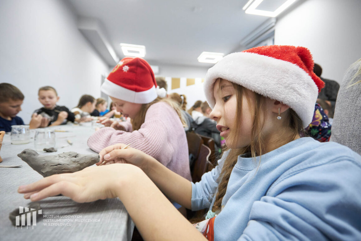 Dwie dziewczynki siedzące przy długim stoliku z innymi dziećmi i lepiące z szarej gliny