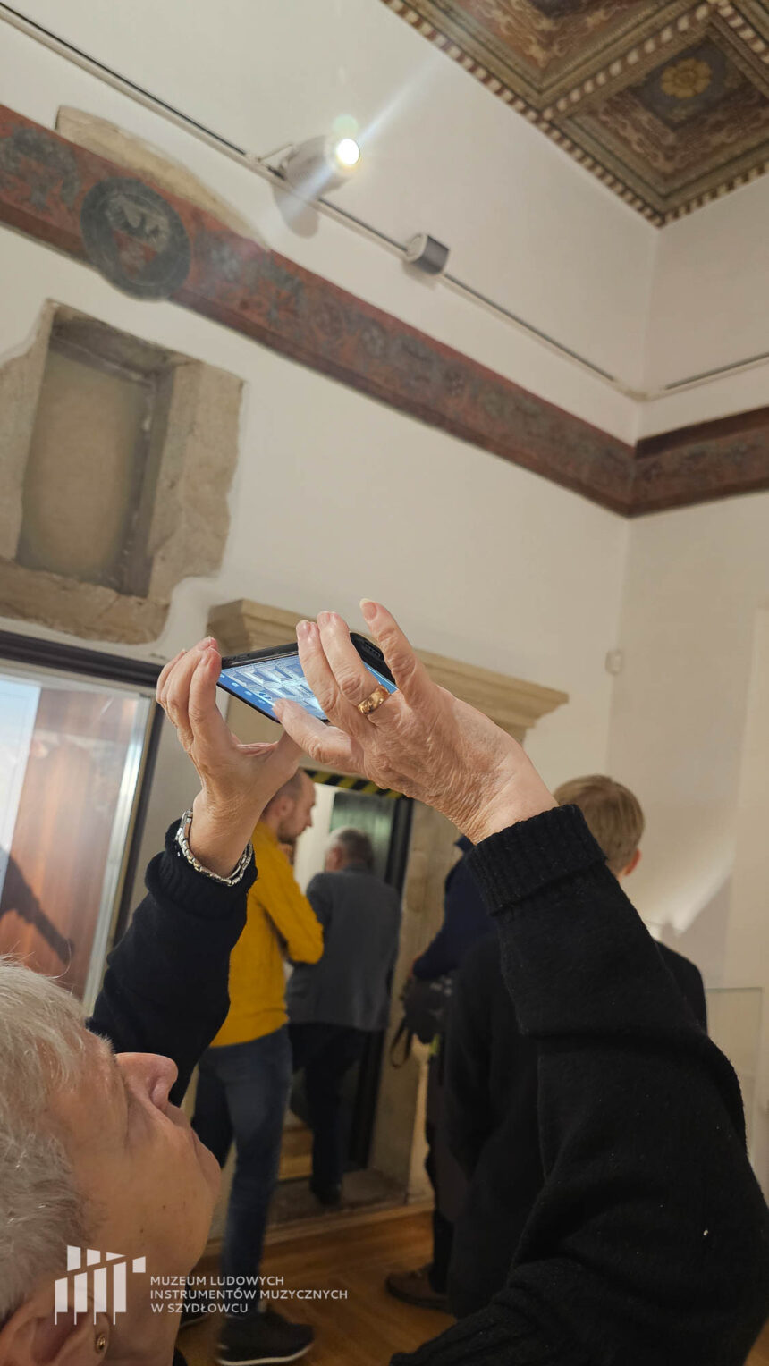 Kobieta robiąca zdjęcie telefonem w muzeum z wysokim malowanym w roślinne wzory drewnianym sufitem z wgłębieniami.